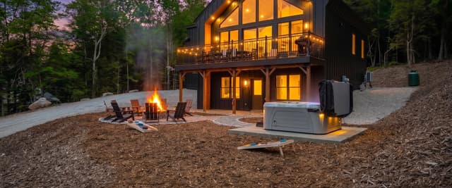 Modern black cabin with glowing windows and deck surrounded by forest, firepit with Adirondack chairs and cornhole in foreground at dusk