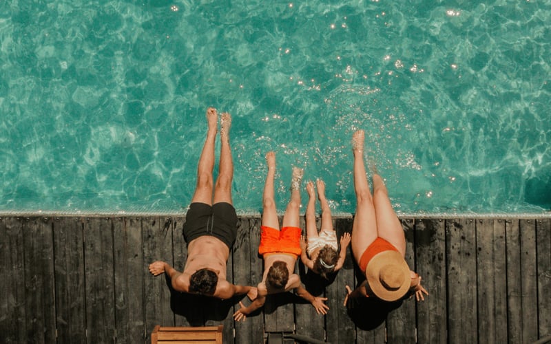 Four people sitting on wooden dock with legs dangling over turquoise water